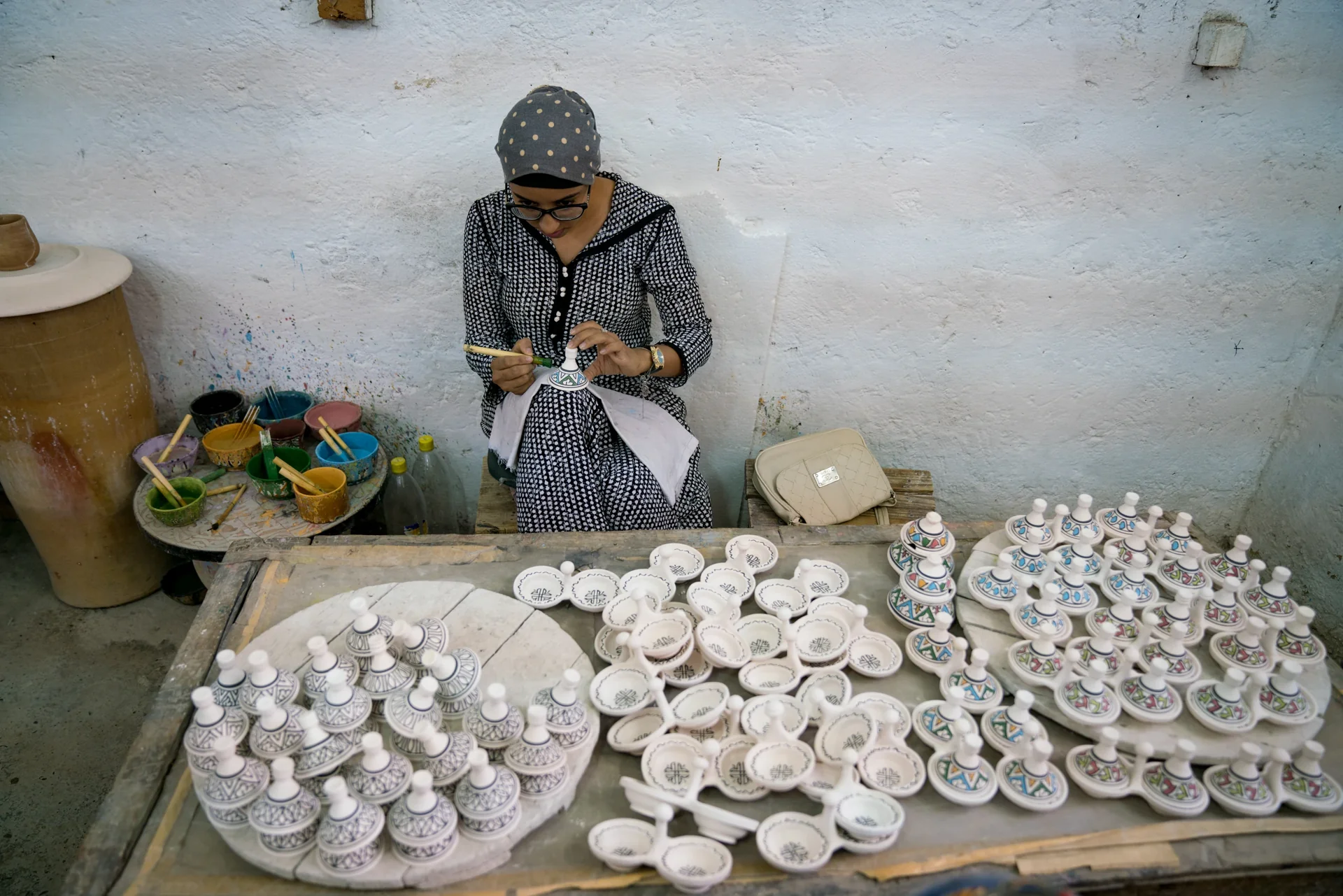 A ceramic craftswoman in Fez paints a small vessel among rows of unfinished white ceramics