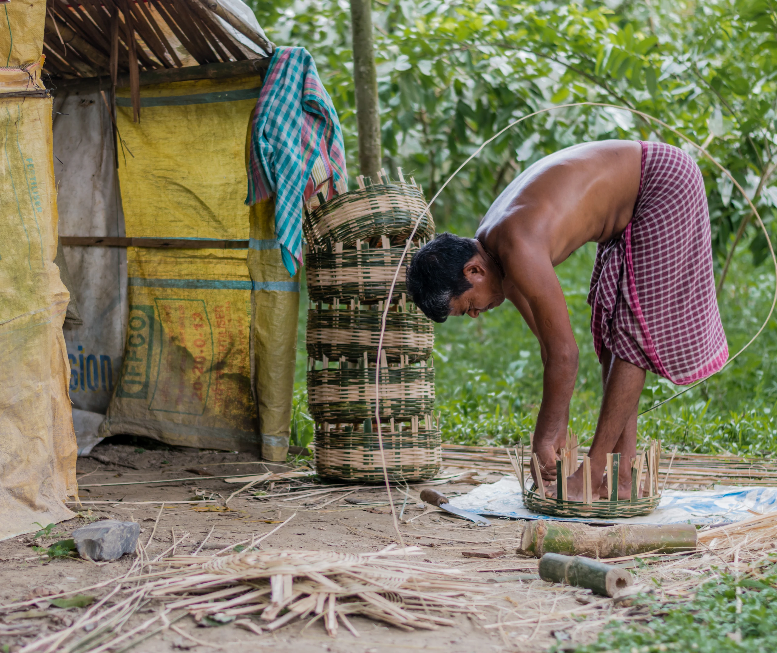 A basket maker bends over a large bamboo basket beside a rural dwelling in India