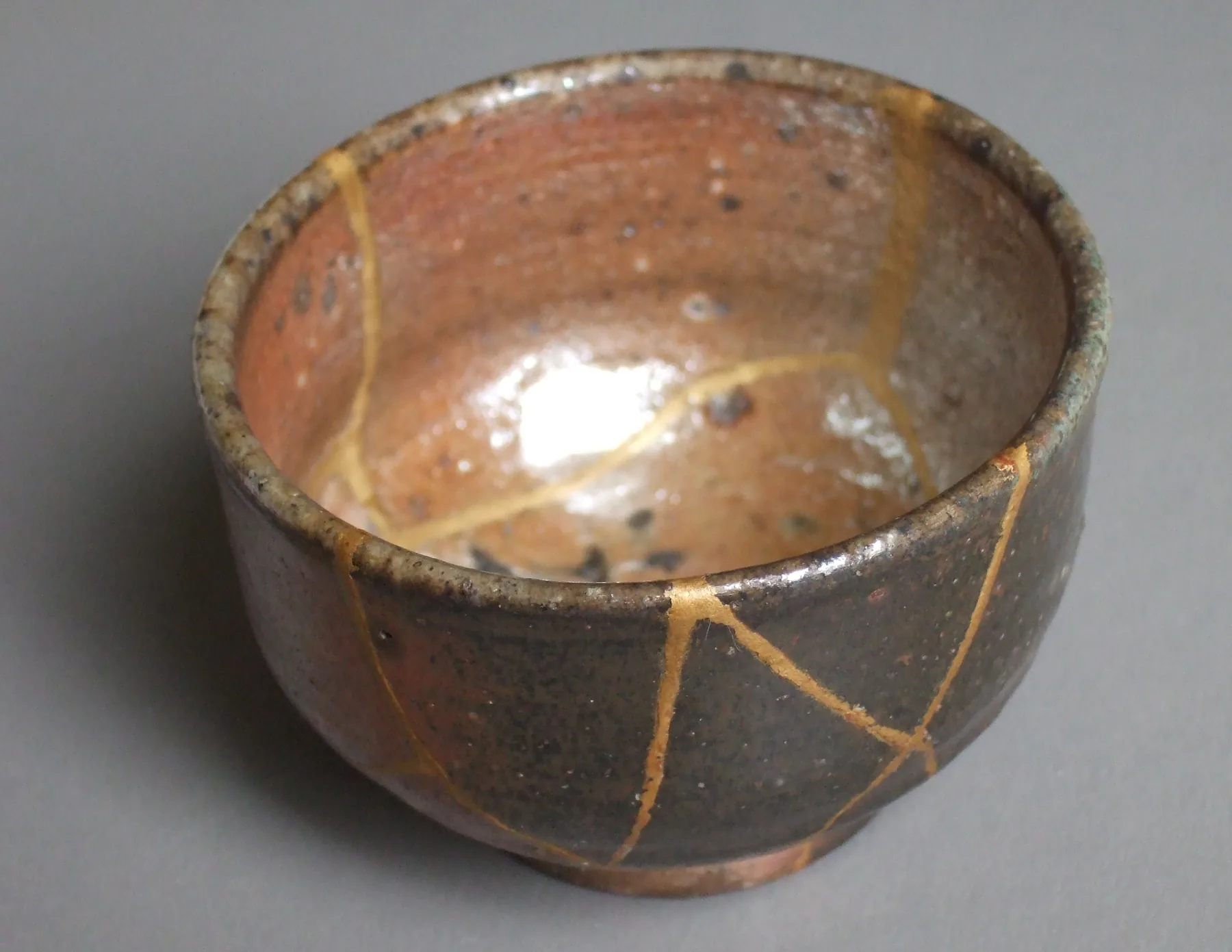 A brown ceramic drinking vessel with gold-coloured repaired cracks visible around its exterior and interior.